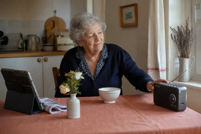 Une femme âgée écoute la radio dans sa cuisine, une tablette posée à proximité.